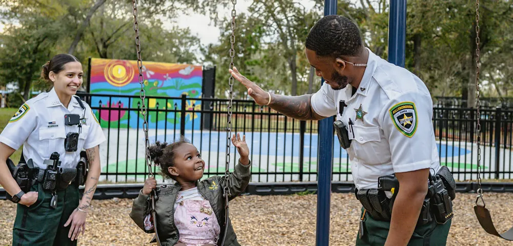 Two Hillsborough County deputies in uniform high-fiving a young girl on a swing at the park