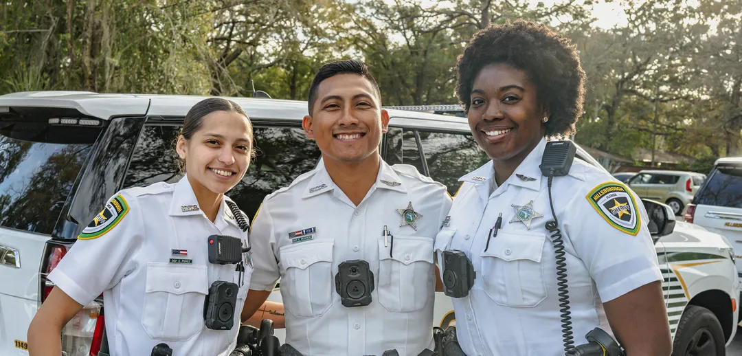 Three smiling patrol deputies in uniform, standing confidently outdoors.